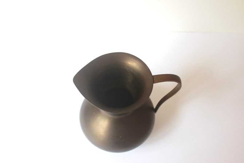 Pode incluir: Um jarro de metal castanho escuro com uma base larga e redonda e uma parte superior em forma de sino. O jarro tem uma &uacute;nica pega.