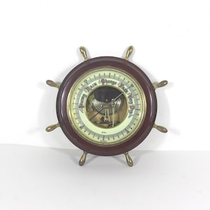 May include: A vintage barometer in the shape of a ship's wheel. The wooden wheel has brass accents and a circular gauge with text indicating weather conditions. The gauge has a cream-colored face with a needle and a glass cover.