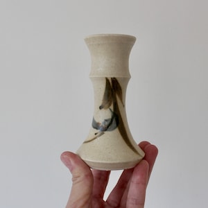 May include: A small, white ceramic vase with a brown and black abstract design. The vase has a narrow neck and a wide base.