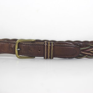 May include: A brown leather belt with a braided design in brown, green, and cream colours. The belt has a brass buckle.