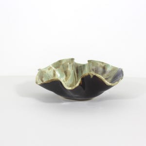 May include: A handmade ceramic bowl with a distinctive, ruffled edge design. The bowl has a dark, glossy black exterior that contrasts with the muted green and tan interior. This decorative bowl is ideal for serving or display.