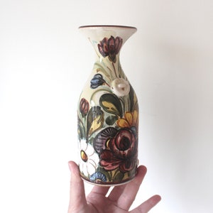 May include: A hand-painted ceramic vase with a cream base and a flared top. The vase features colourful floral designs in red, purple, yellow, and white, with green leaves. It has a small handle.