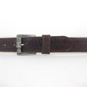 May include: A brown leather belt with a silver buckle. The belt has a textured surface and a pointed end.