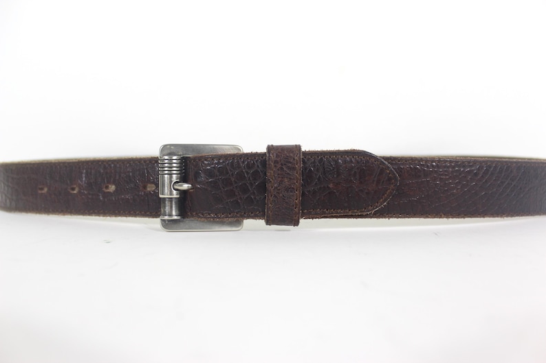 May include: A brown leather belt with a silver buckle. The belt has a textured surface and a pointed end.
