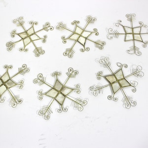 May include: Six decorative snowflake ornaments. Each snowflake has a central square with a light-coloured inlay, possibly mother-of-pearl. The delicate silver-toned metalwork has curled accents and arrow-like points.