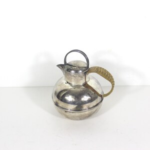 Peut inclure: Une théière sphérique argentée avec une poignée tressée et un petit couvercle rond. La théière a un bec verseur et une poignée noire sur le couvercle. La poignée est en matériau tressé brun clair.