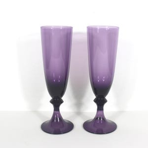 May include: Two purple glass champagne flutes with a gradient from light to dark purple. The flutes have a rounded bowl and a stem with a small knob at the base.