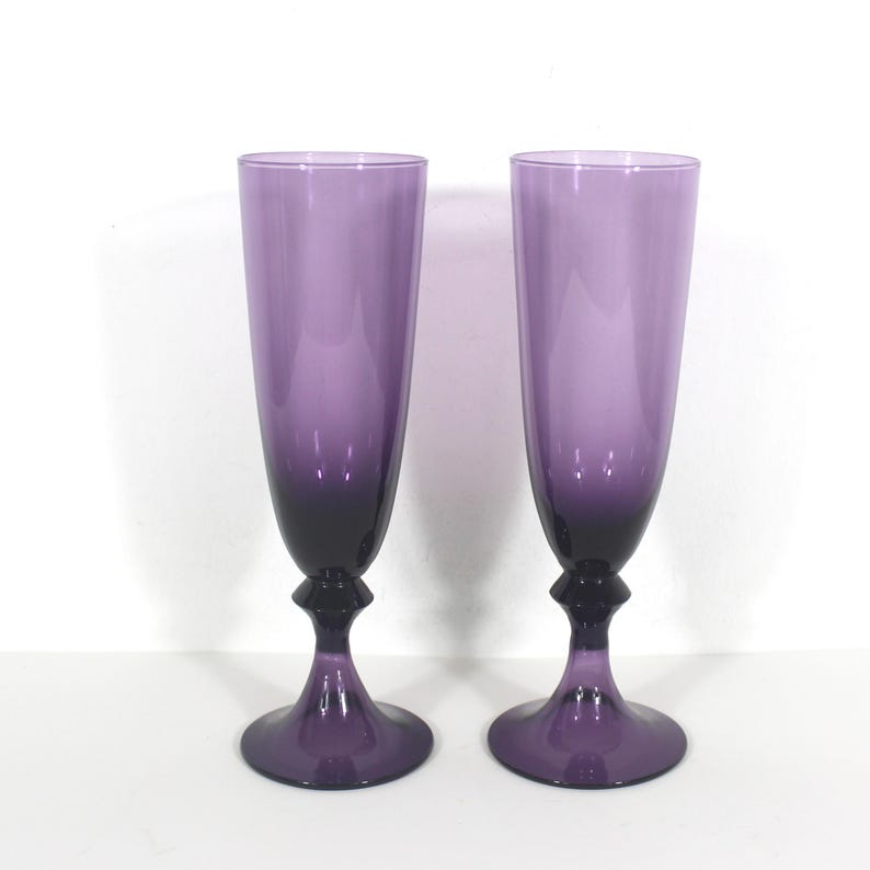 May include: Two purple glass champagne flutes with a gradient from light to dark purple. The flutes have a rounded bowl and a stem with a small knob at the base.