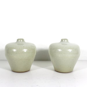 May include: Two ceramic vases in a light green color with a smooth, slightly textured finish. The vases have a rounded, bulbous shape with a narrow opening at the top.