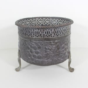 May include: A vintage metal planter with a decorative grape design and a pierced top rim. The planter has three legs and is made of a dark grey metal.