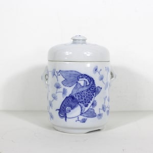 May include: A white ceramic jar with a blue and white design of a fish and floral pattern. The jar has a lid and two handles.