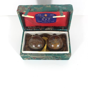 May include: Two dark brown spherical metal balls with gold-colored dragon designs inside a rectangular green fabric-covered box. The box lid interior is red with a blue label featuring the text "HEALTHY BALL" and Chinese characters.