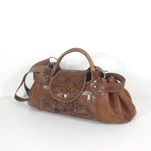 May include: A brown leather handbag featuring an embossed floral pattern on the front flap and body. The bag includes a top handle, a detachable shoulder strap, silver-toned metal hardware, and a zippered closure with a metallic pull tab.