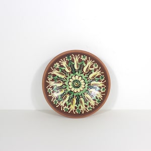 May include: A round, brown ceramic plate with a floral design. The plate features a central flower motif surrounded by green and yellow stylized floral elements. The rim is a darker brown.