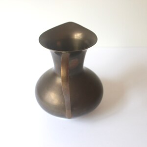 Pode incluir: Um jarro de metal castanho escuro com uma pega curva. O jarro tem um corpo redondo e uma abertura larga e em forma de sino.