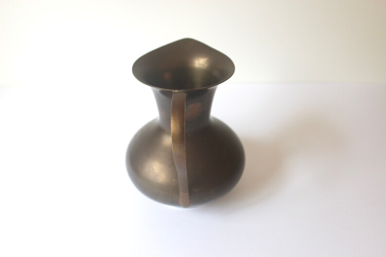Pode incluir: Um jarro de metal castanho escuro com uma pega curva. O jarro tem um corpo redondo e uma abertura larga e em forma de sino.