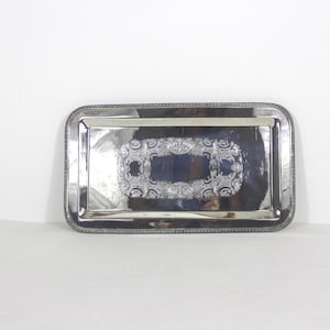 May include: A rectangular silver serving tray with a decorative border and a central ornate design. The tray has a polished, reflective surface and a raised edge. The central design features a rectangular shape with floral and scrollwork details.