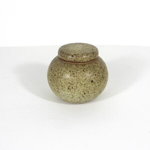 May include: A small, round ceramic jar with a matching lid, featuring a speckled light green and tan glaze. The container has a bulbous body and a smooth, rounded top, showcasing a natural, earthy texture with dark brown flecks throughout.