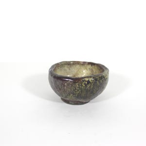 May include: A small, handcrafted ceramic bowl with a rough, textured exterior. The bowl features a dark brown rim and a mottled green and brown body, with a lighter interior. The bowl is approximately 3 inches in diameter, and is suitable for serving small portions.