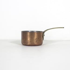 May include: A small copper saucepan with a brass handle. The pot has a rounded body and a silver interior. The handle curves upwards. The saucepan is likely used for cooking small portions. The handle is made of brass.