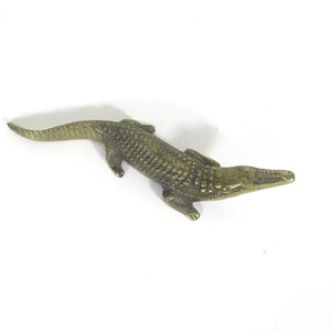 May include: A detailed, bronze-colored alligator figurine. The reptile is lying down, with its textured skin and scales clearly visible. The figurine is a decorative item, likely for display.