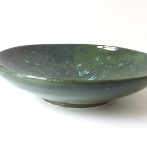 May include: A green ceramic bowl with a speckled glaze. The bowl has a wide, shallow shape and a slightly raised rim.