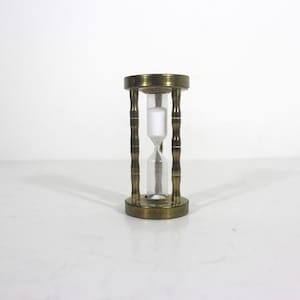 May include: A brass hourglass with a clear glass bulb and white sand. The hourglass is on a white surface.