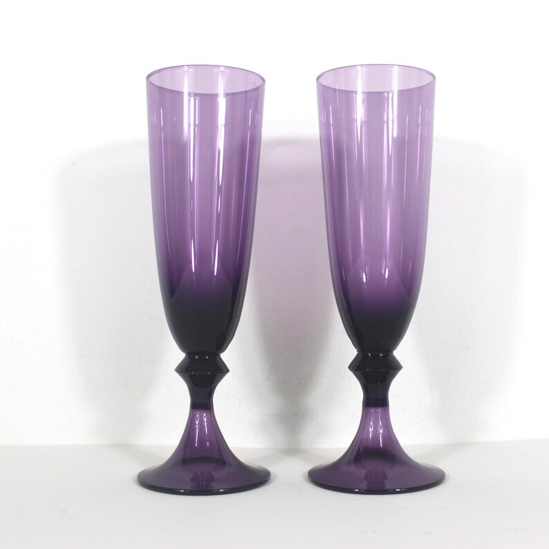 May include: Two purple glass champagne flutes with a gradient from dark to light purple. The flutes have a unique, flared base.
