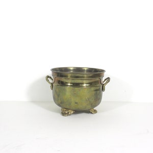 May include: A brass planter with three feet and two handles. The planter is a light gold colour.