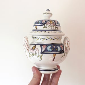 May include: A white ceramic jar with a lid, decorated with blue, green and pink floral patterns. The jar has two handles and a round base.