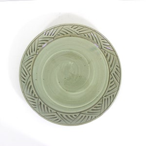 May include: A round, pale green ceramic plate with a decorative border. The border features a repeating geometric pattern. The plate's centre has a spiral design. The plate is set against a white background.