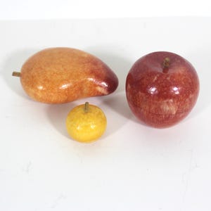 May include: Three decorative fruit sculptures made of marble. The fruit are a mango, an apple, and a small lemon. The mango is orange and yellow, the apple is red, and the lemon is yellow.