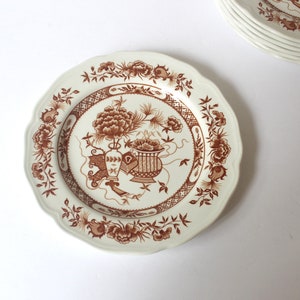 May include: A white ceramic dinner plate with a brown floral design. The design features stylised flowers, leaves, and vases.