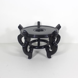 May include: A black, ornate wooden stand with a round top and decorative curved legs. The stand has a glossy finish and a circular opening in the centre of the top. The stand is designed to hold a decorative object.