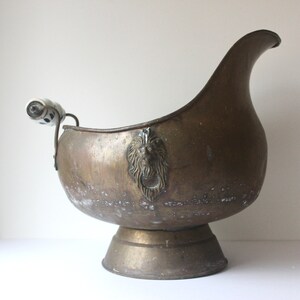 May include: An antique brass ice bucket with a lion head design and a white porcelain handle. The bucket has a rounded base and a wide, curved opening.