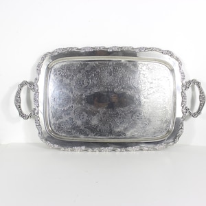 May include: A rectangular silver-toned serving tray featuring an ornate, embossed floral and scrollwork border. The tray includes two matching decorative handles on the sides and a central surface area decorated with intricate, etched botanical patterns.