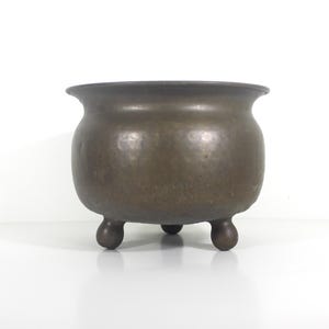 May include: A bronze-coloured, three-legged cauldron with a rounded body and a flared rim. The metal pot has a hammered texture and a dark, aged patina, suggesting it is an antique or vintage piece. The cauldron is designed to sit on a flat surface.