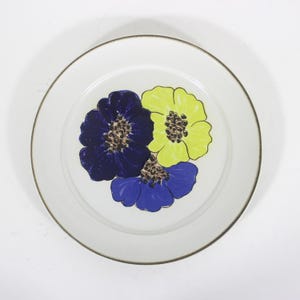 May include: A white ceramic dinner plate with a brown rim features a stylised floral design. Three flowers are depicted in shades of blue and yellow.