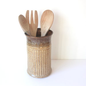 May include: A brown and beige ceramic utensil holder with a ribbed design, holding three wooden kitchen utensils: a spoon, a spatula, and a fork.