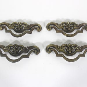 May include: Four antique brass drawer pulls with ornate scrollwork and a dark patina. Each pull has a central decorative element and curved handles. The pulls are arranged on a white surface.