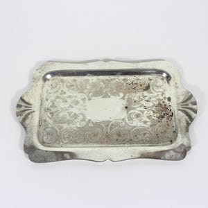 Peut inclure: Un plateau rectangulaire en métal argenté présentant un motif ornemental en relief sur toute la surface. Le plateau possède un bord surélevé aux contours festonnés et des motifs en éventail sur les côtés, avec des traces d'oxydation.