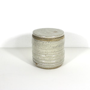 May include: A cylindrical ceramic canister with a lid, featuring a speckled off-white glaze and horizontal ribbed texture. The base and rim of the lid show exposed brown clay, and the surface has small dark flecks throughout the finish.