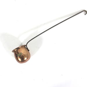 May include: A copper ladle with a long, thin handle. The ladle has a rounded bowl and a small, rectangular label on the side.