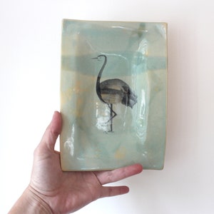 May include: Rectangular ceramic tray with a hand-painted crane design. The tray has a light blue and yellow background with a black and grey crane illustration. The tray is held by a hand.