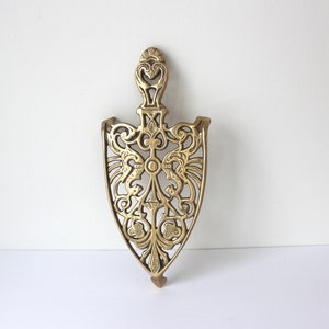 May include: A gold-toned metal trivet with an intricate, ornate design. The trivet is shaped like a shield with a pointed top and a rounded bottom.