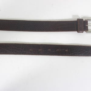 May include: A brown leather belt with a silver buckle. The belt is made of a textured leather and has a simple design.