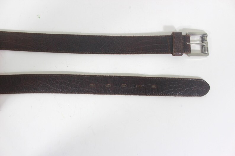 May include: A brown leather belt with a silver buckle. The belt is made of a textured leather and has a simple design.