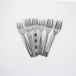 May include: Six stainless steel forks with decorative handle designs. The forks are arranged in a fan shape against a white background. The handles feature various patterns, including floral and geometric motifs, adding a touch of elegance to the silverware.