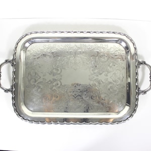 May include: A rectangular silver-toned serving tray featuring an ornate, engraved floral pattern across its centre. The tray has a scalloped edge and two decorative handles on opposite sides, each accented with a small shell-like motif at the base.