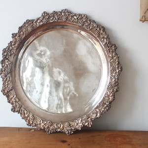 May include: A silver serving tray with a decorative grapevine border. The tray is round and has a smooth, reflective surface.